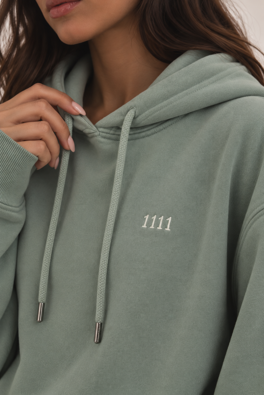 Close-up of a person wearing a sage green pullover hoodie with white drawstrings and a small white '1111' embroidered on the chest.