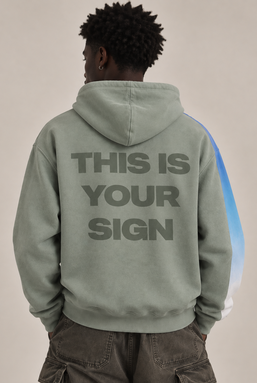 A person shown from the back wearing a sage green hoodie with the text 'THIS IS YOUR SIGN' printed in dark gray across the back, paired with dark cargo pants against a plain beige studio background.