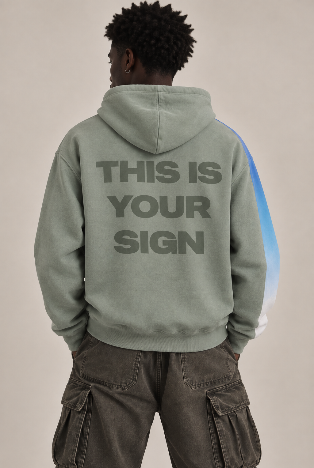 A person shown from the back wearing a sage green hoodie with the text 'THIS IS YOUR SIGN' printed in dark gray across the back, paired with dark cargo pants against a plain beige studio background.