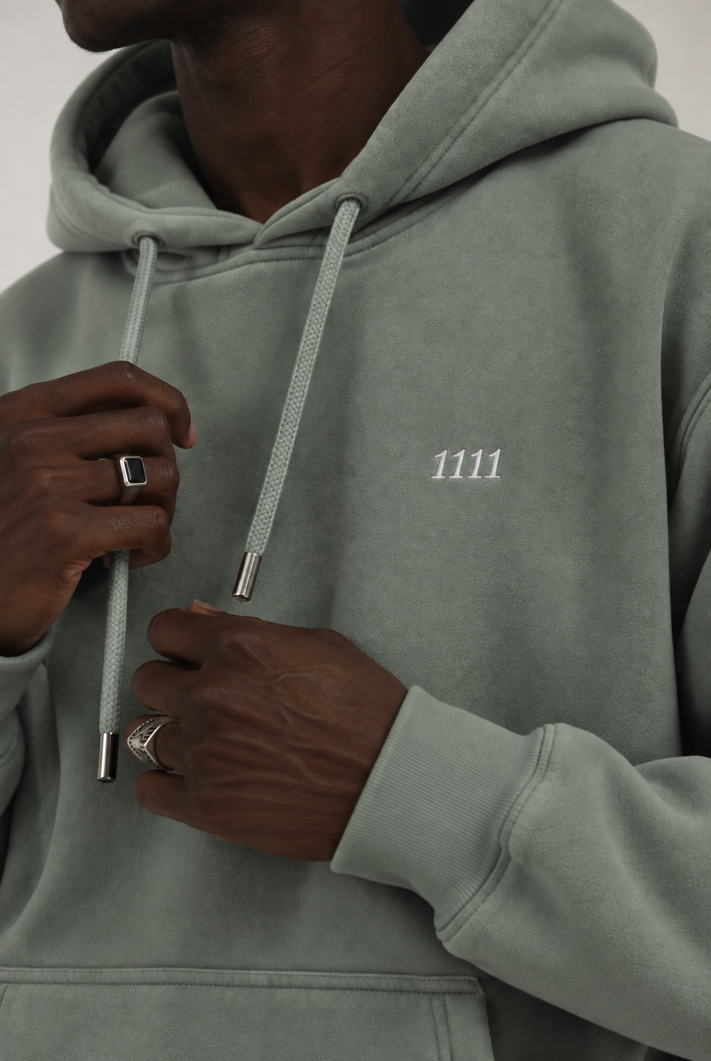 A close-up of a person wearing a sage green hooded sweatshirt with white drawstrings and a small white '1111' logo on the chest.