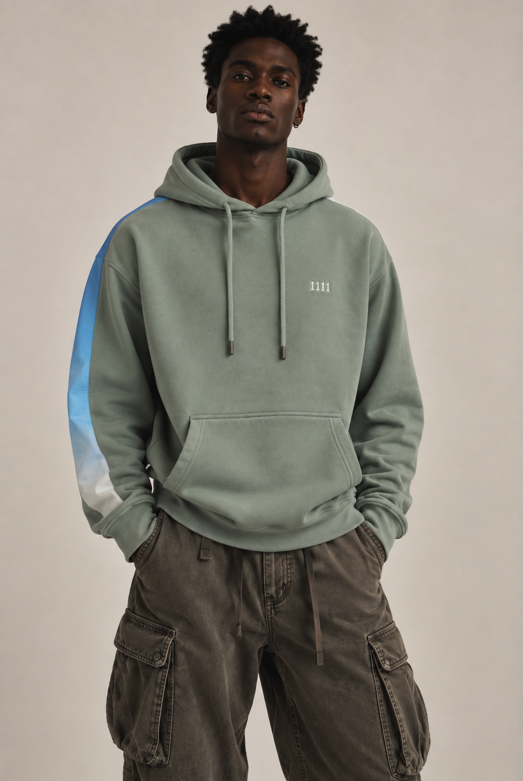 A man wearing a sage green pullover hoodie with a light blue and white gradient stripe down the sleeve, and a white embroidered '1111' on the left chest, paired with dark brown cargo pants, against a plain beige background.