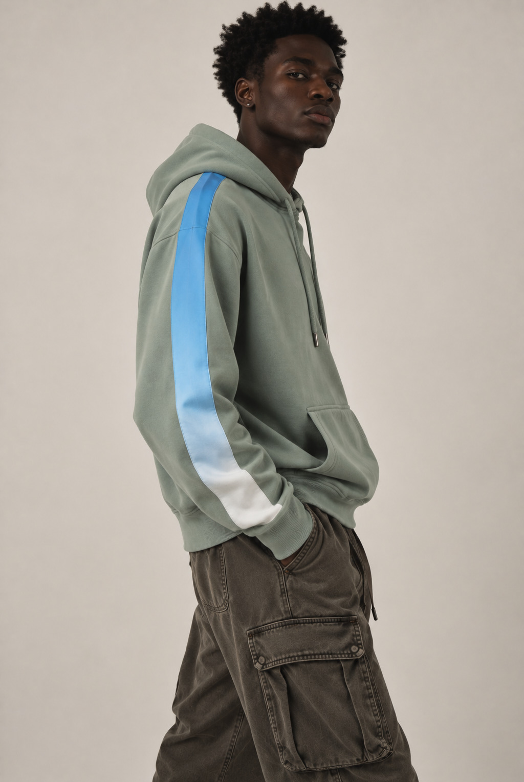 A man wearing a sage green hoodie with a light blue and white gradient stripe down the sleeve, paired with dark brown cargo pants, photographed against a neutral light gray background.