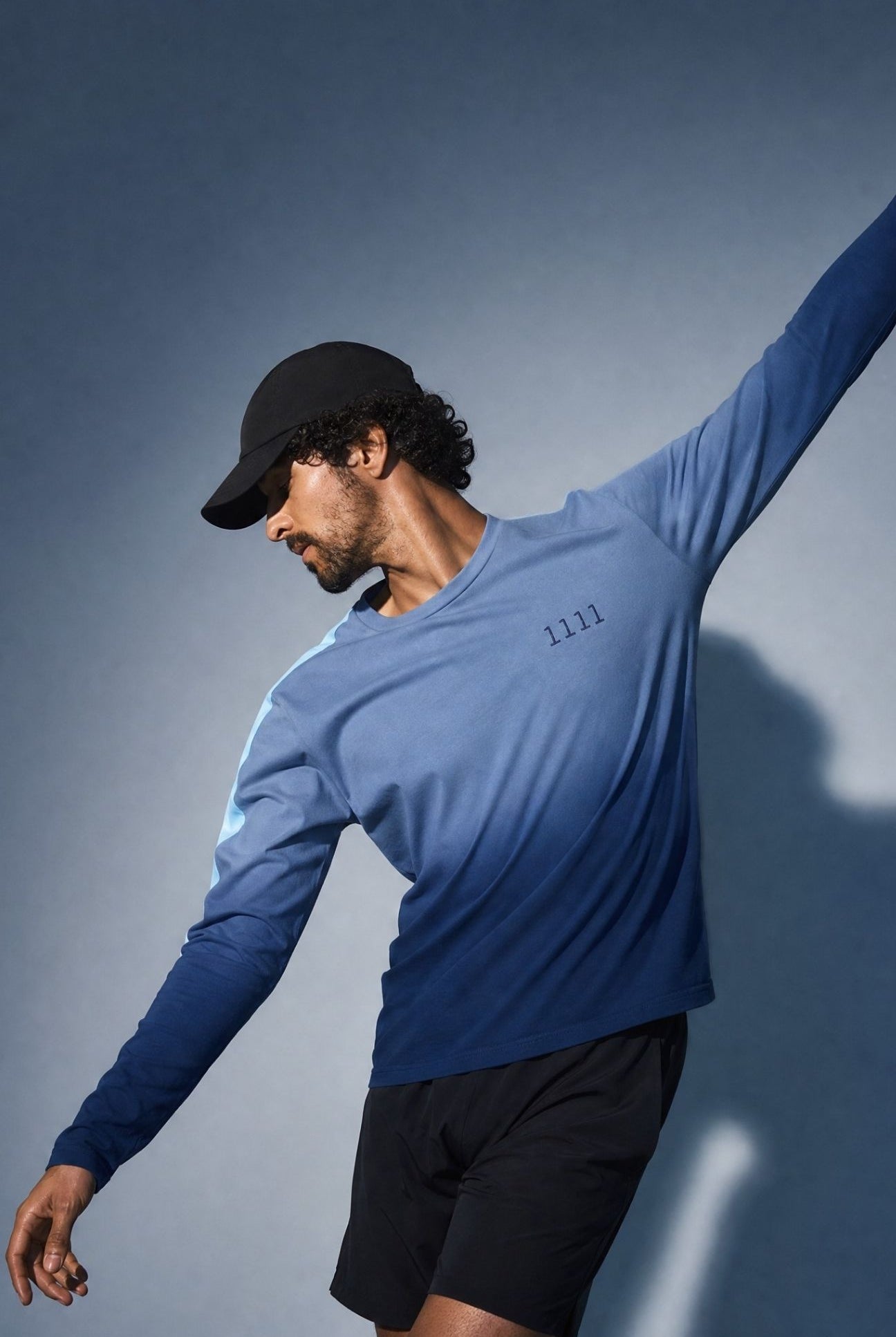 A man wearing a blue long-sleeve shirt with a gradient design and an embroidered 1111 in navy, black shorts, and a black baseball cap, posing dynamically against a dark gray studio background.
