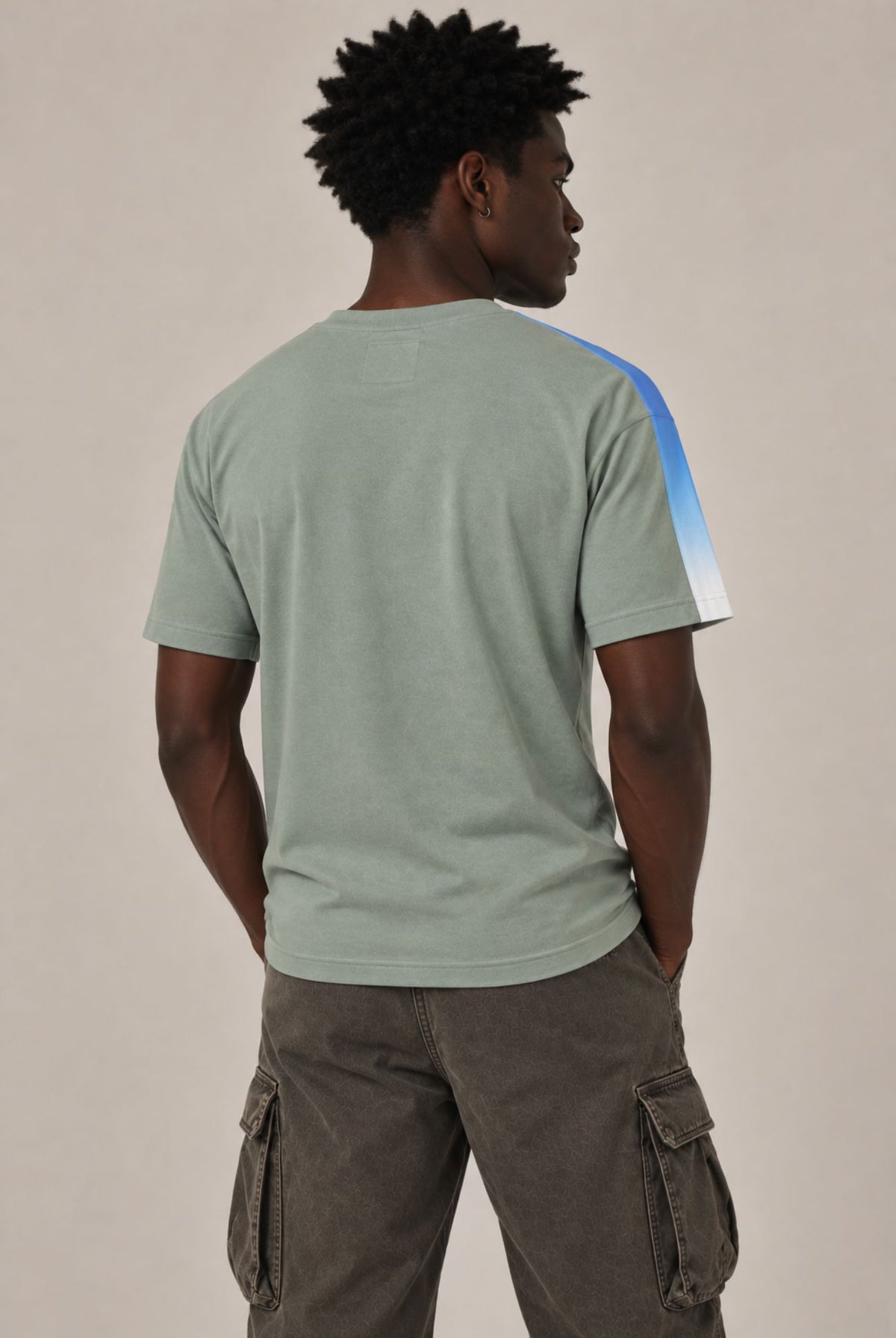 A man viewed from behind wearing a sage green t-shirt with a blue and white color-blocked stripe on the right shoulder, paired with brown cargo pants, against a plain beige background.