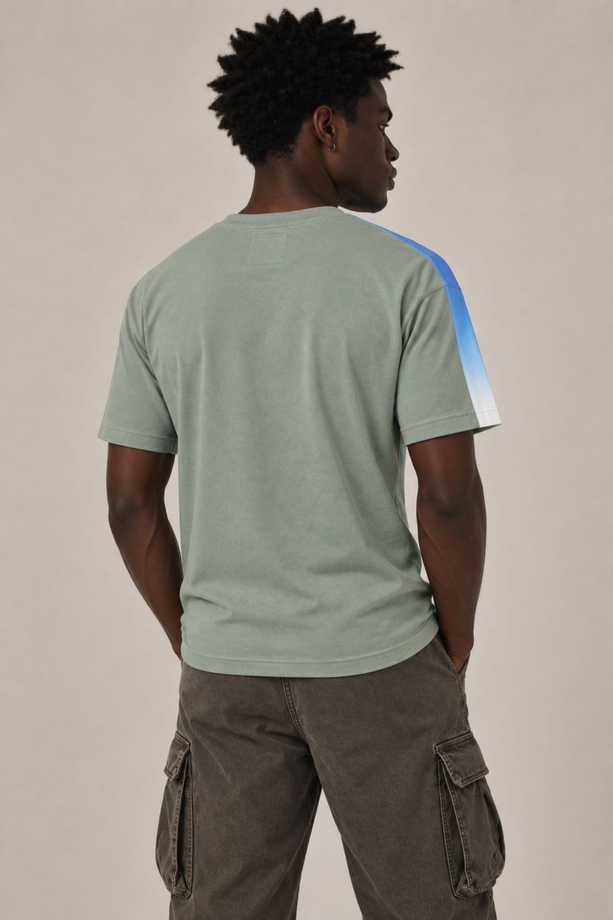 A man viewed from behind wearing a sage green t-shirt with a blue and white color-blocked stripe on the right shoulder, paired with brown cargo pants, against a plain beige background.
