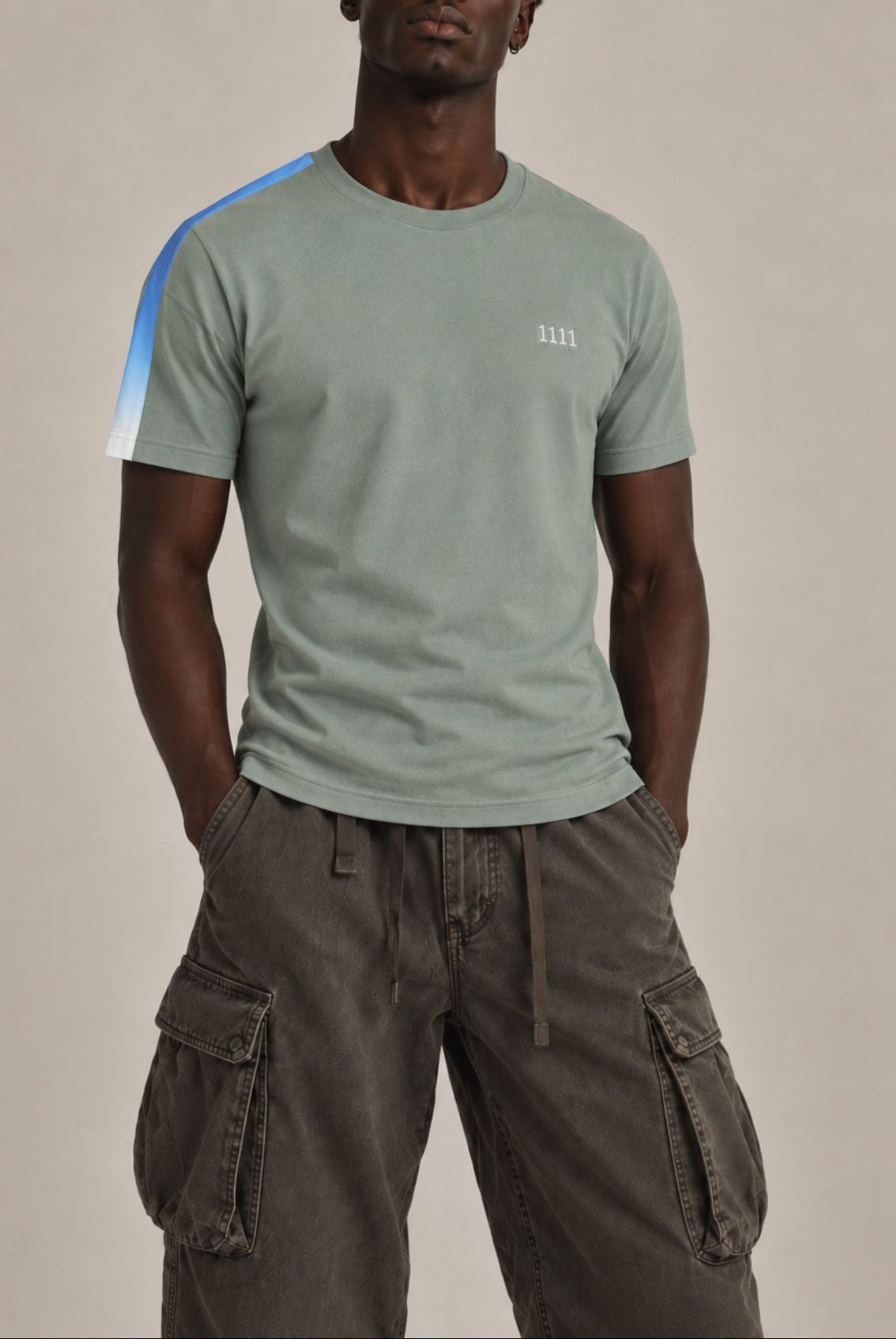 A man wearing a sage green crewneck t-shirt with a blue and white diagonal stripe on the right sleeve and a small white embroidered 1111 on the left chest, paired with dark brown cargo pants, against a solid beige studio background.