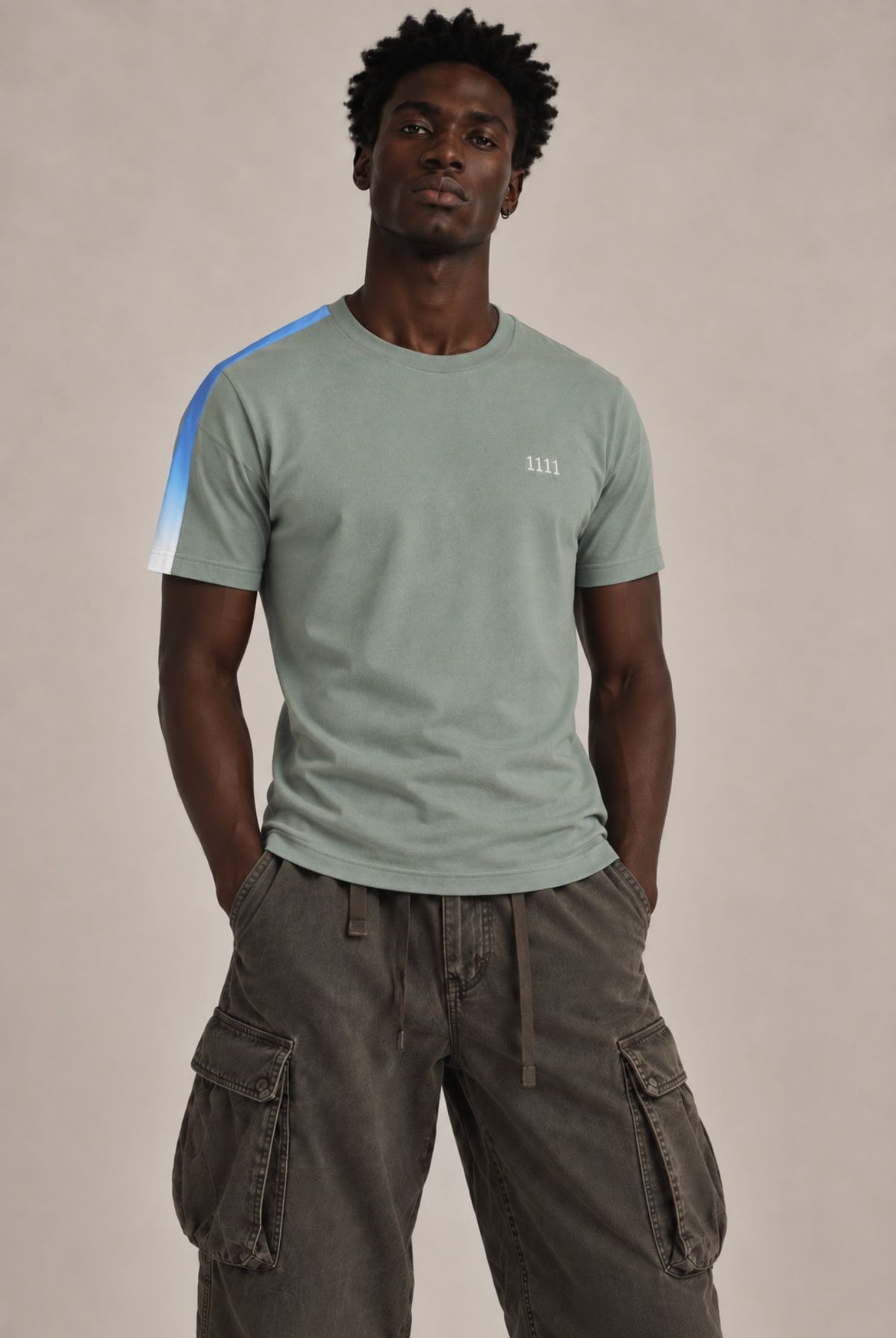 A man wearing a sage green crewneck t-shirt with a blue and white diagonal stripe on the right sleeve and a small white embroidered 1111 on the left chest, paired with dark brown cargo pants, against a solid beige studio background.