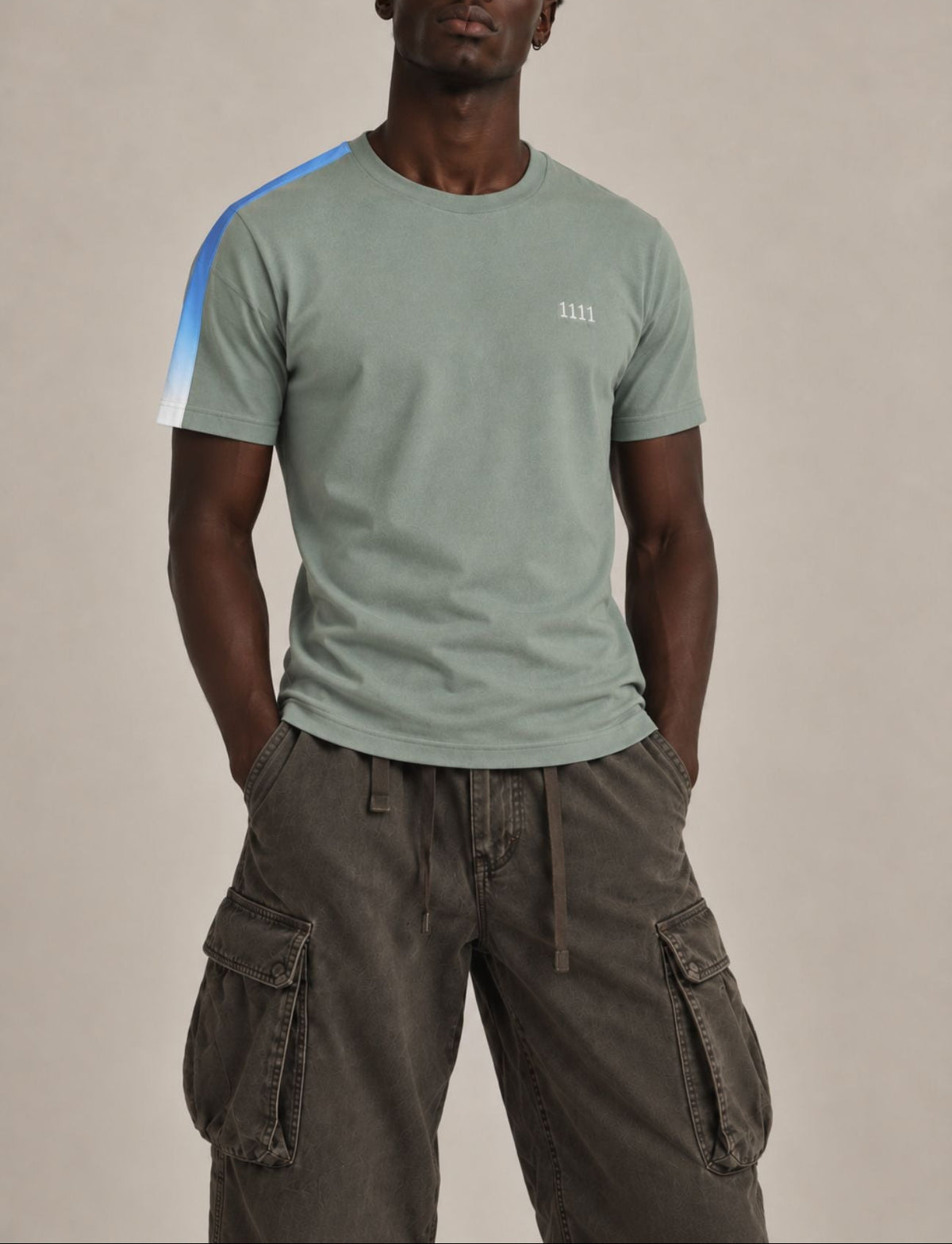 A man wearing a sage green crewneck t-shirt with a blue and white diagonal stripe on the right sleeve and a small white embroidered 1111 on the left chest, paired with dark brown cargo pants, against a solid beige studio background.