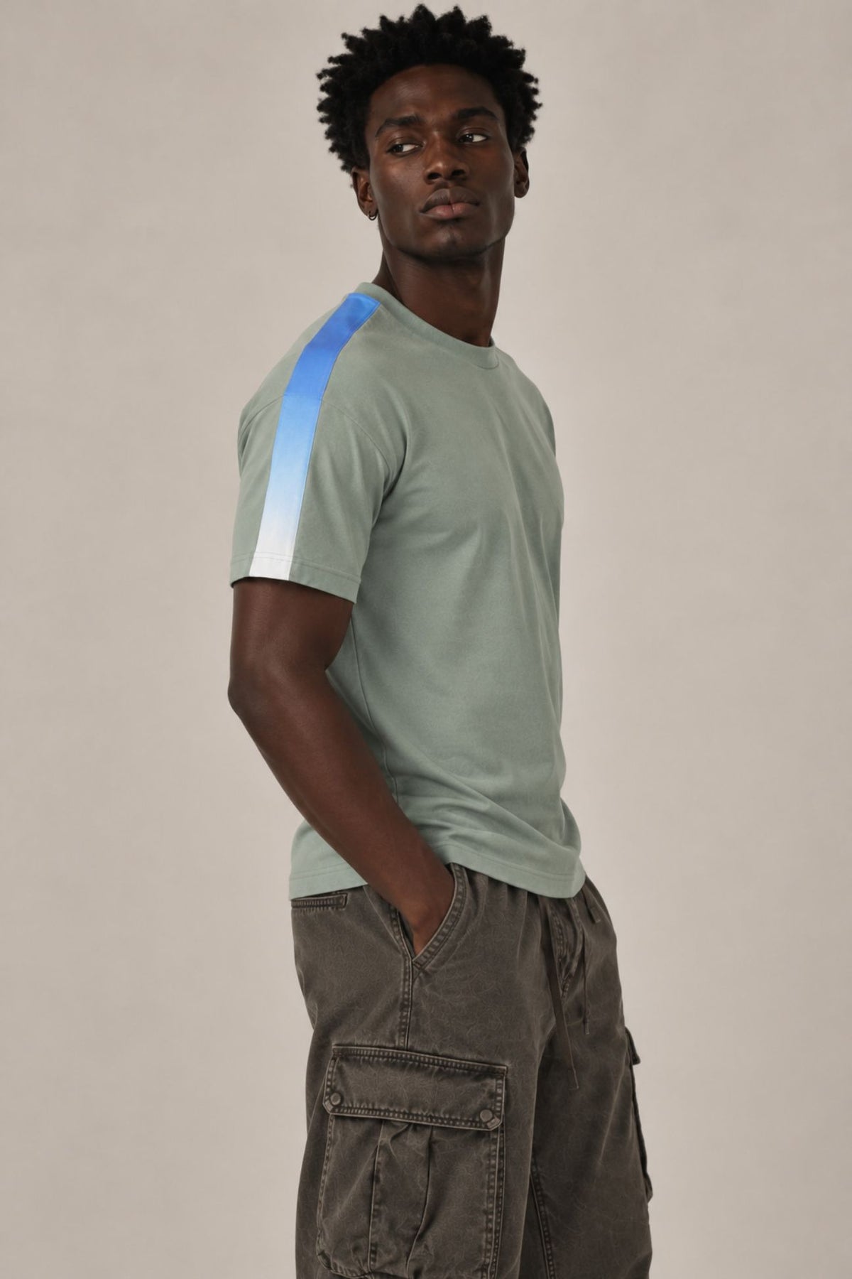 Side view of male model wearing a sage green crewneck t-shirt with white embroidered 1111 angel number detail and a blue-to-white gradient stripe along the right shoulder and sleeve, paired with dark gray cargo pants, photographed against a neutral beige studio background.
