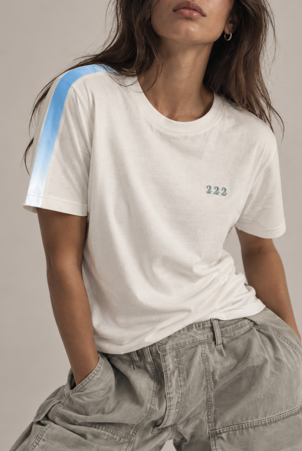 A woman wearing a white short-sleeve crewneck t-shirt with a light blue gradient stripe on the right shoulder and the number '222' embroidered in green on the left chest, paired with gray cargo pants.