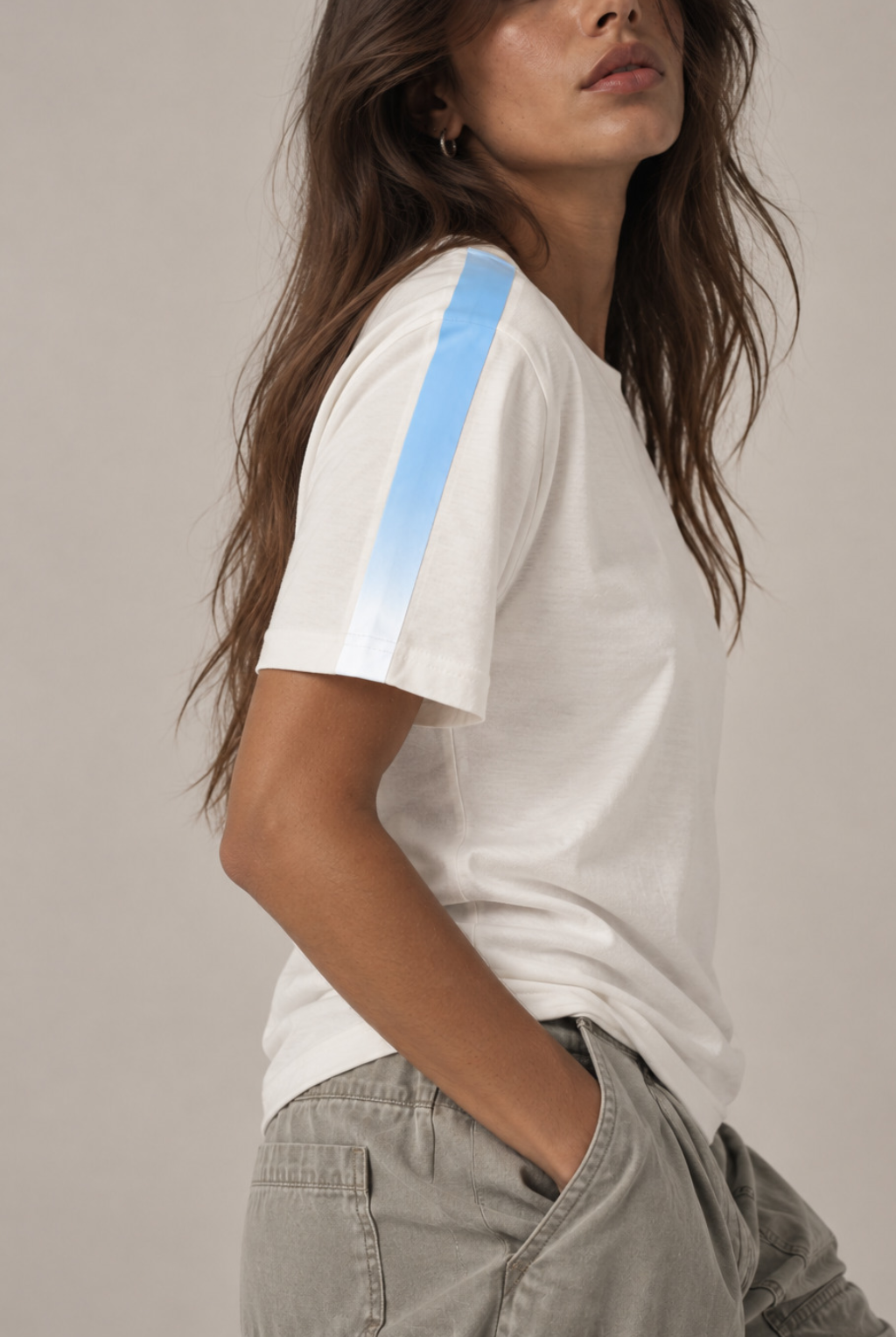 A woman wearing a white short-sleeve crewneck t-shirt with a light blue gradient stripe down the right sleeve, paired with light gray cargo pants, set against a plain light gray background.