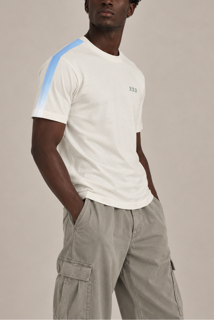 A man wearing a white short-sleeve t-shirt with a light blue stripe detail on the right shoulder and a small green '222' embroidery on the chest, paired with light gray cargo pants, against a solid light gray background.