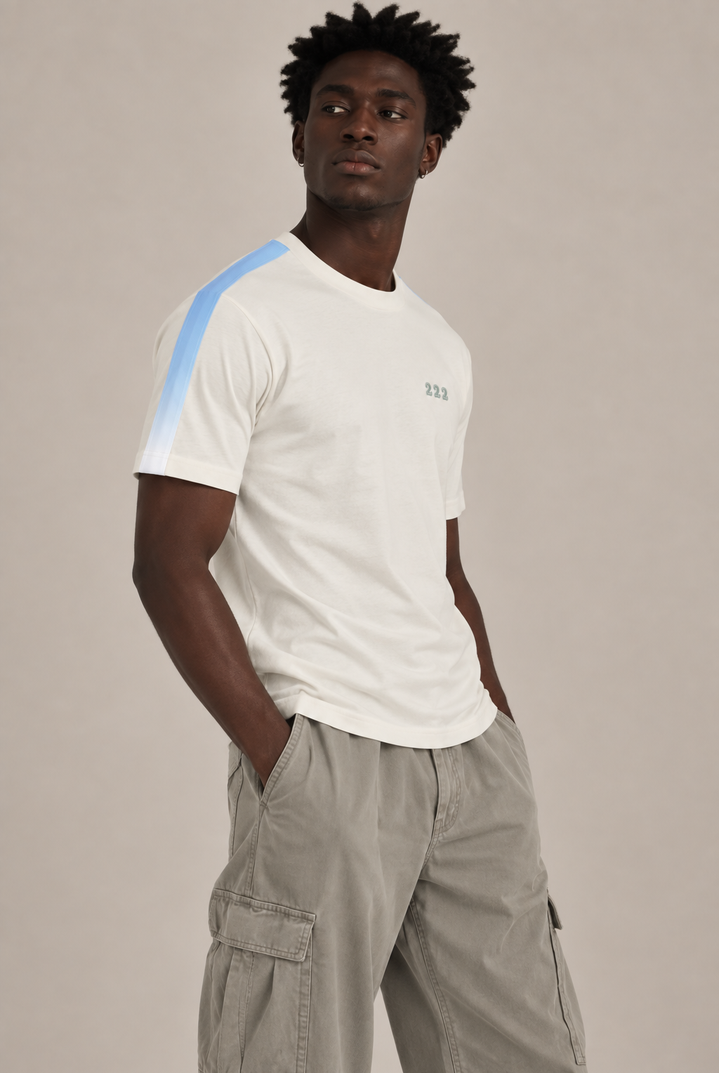 A man wearing a white short-sleeve t-shirt with a light blue stripe detail on the right shoulder and a small green '222' embroidery on the chest, paired with light gray cargo pants, against a solid light gray background.