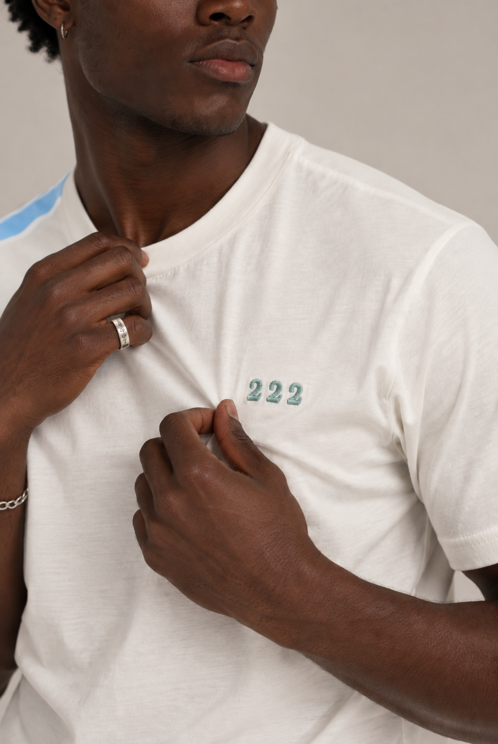A close-up of a man wearing a white crew-neck t-shirt with a small green '222' embroidery on the chest. He is adjusting the collar with both hands, showing a silver ring on one finger and a chain bracelet on the wrist.