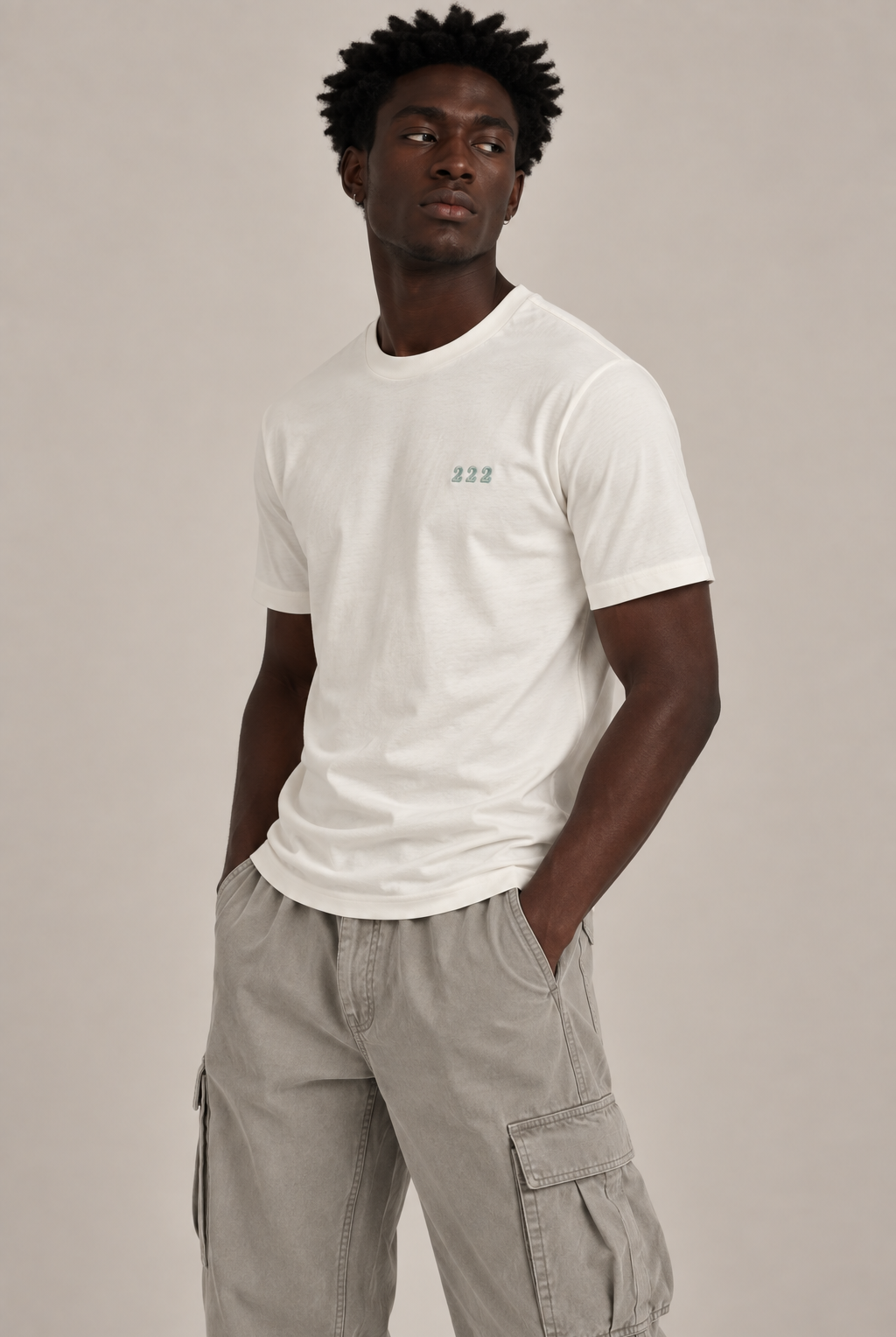 A man wearing a white crew-neck t-shirt with a small green '222' graphic on the left chest, paired with light gray cargo pants, against a solid light gray background.