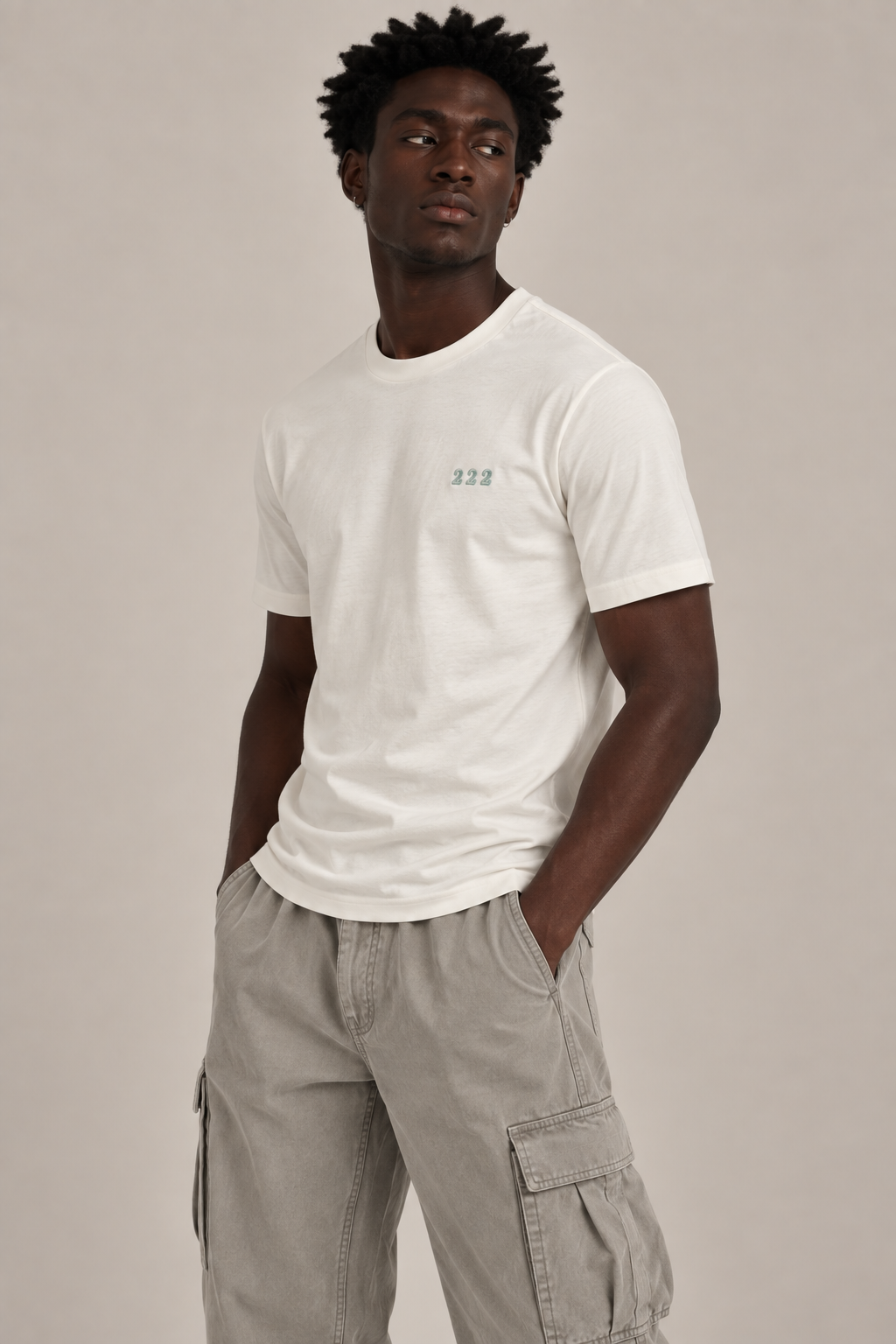 A man wearing a white crew-neck t-shirt with a small green '222' graphic on the left chest, paired with light gray cargo pants, against a solid light gray background.