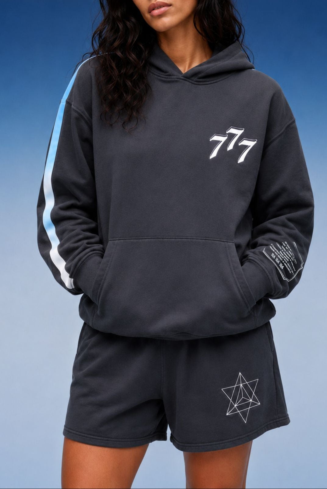 A woman modeling a dark gray hoodie and matching shorts set against a blue gradient background. The hoodie features a white '777' graphic on the chest, a light blue and white stripe down one sleeve, and a white geometric star graphic on the shorts.