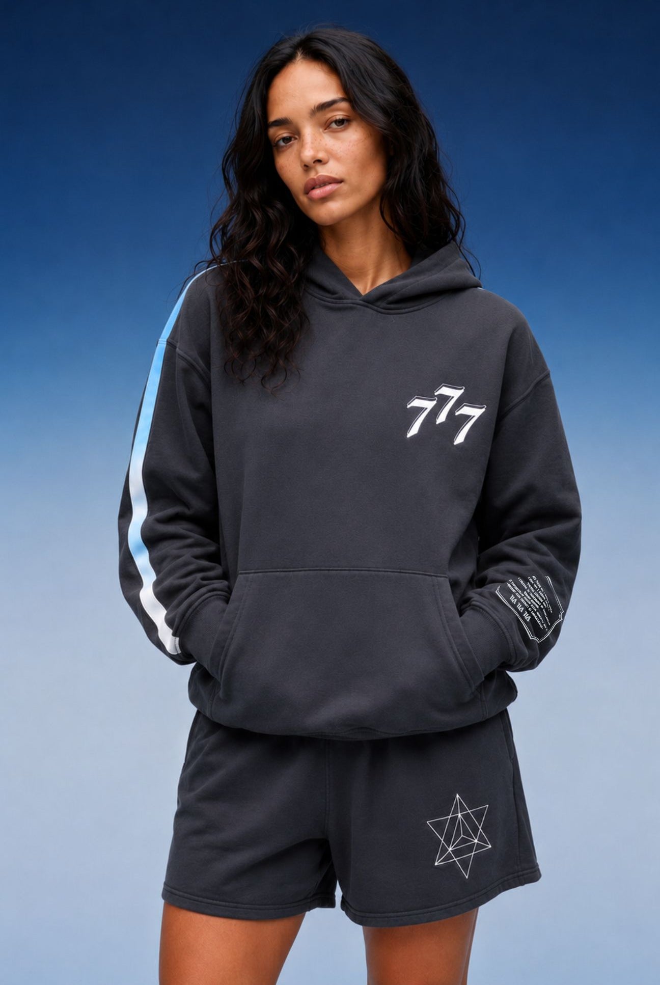 A woman modeling a dark gray hoodie and matching shorts set against a blue gradient background. The hoodie features a white '777' graphic on the chest, a light blue and white stripe down one sleeve, and a white geometric star graphic on the shorts.