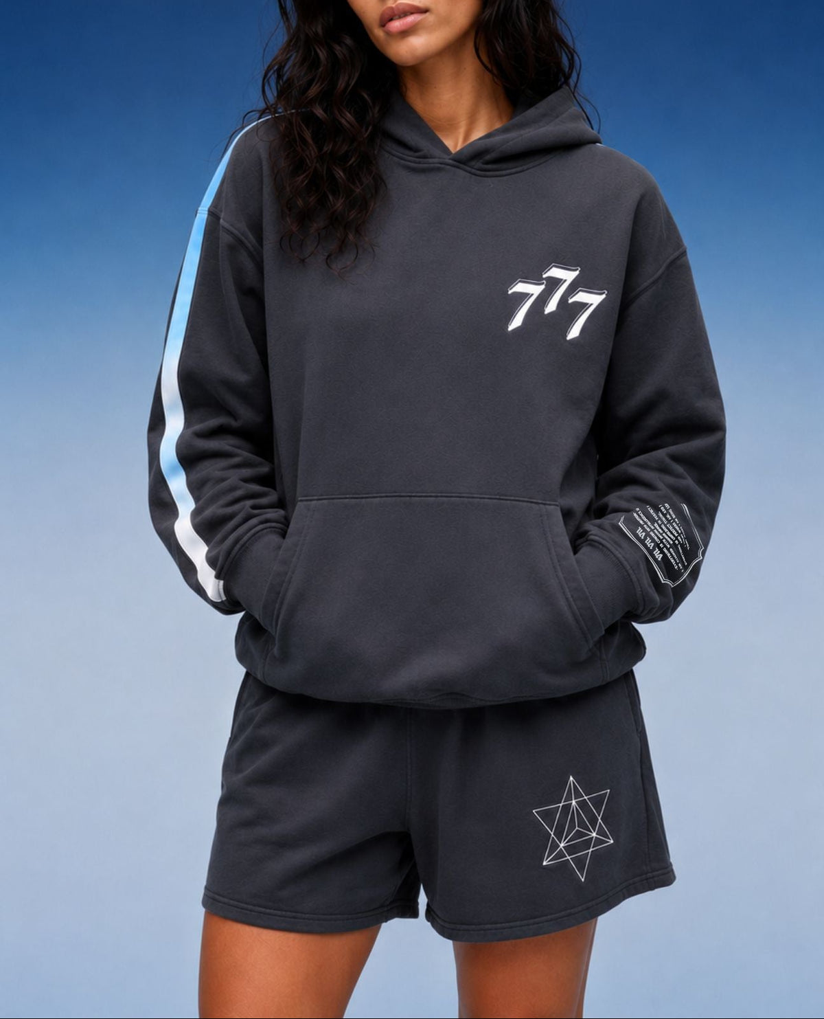 A woman modeling a dark gray hoodie and matching shorts set against a blue gradient background. The hoodie features a white '777' graphic on the chest, a light blue and white stripe down one sleeve, and a white geometric star graphic on the shorts.