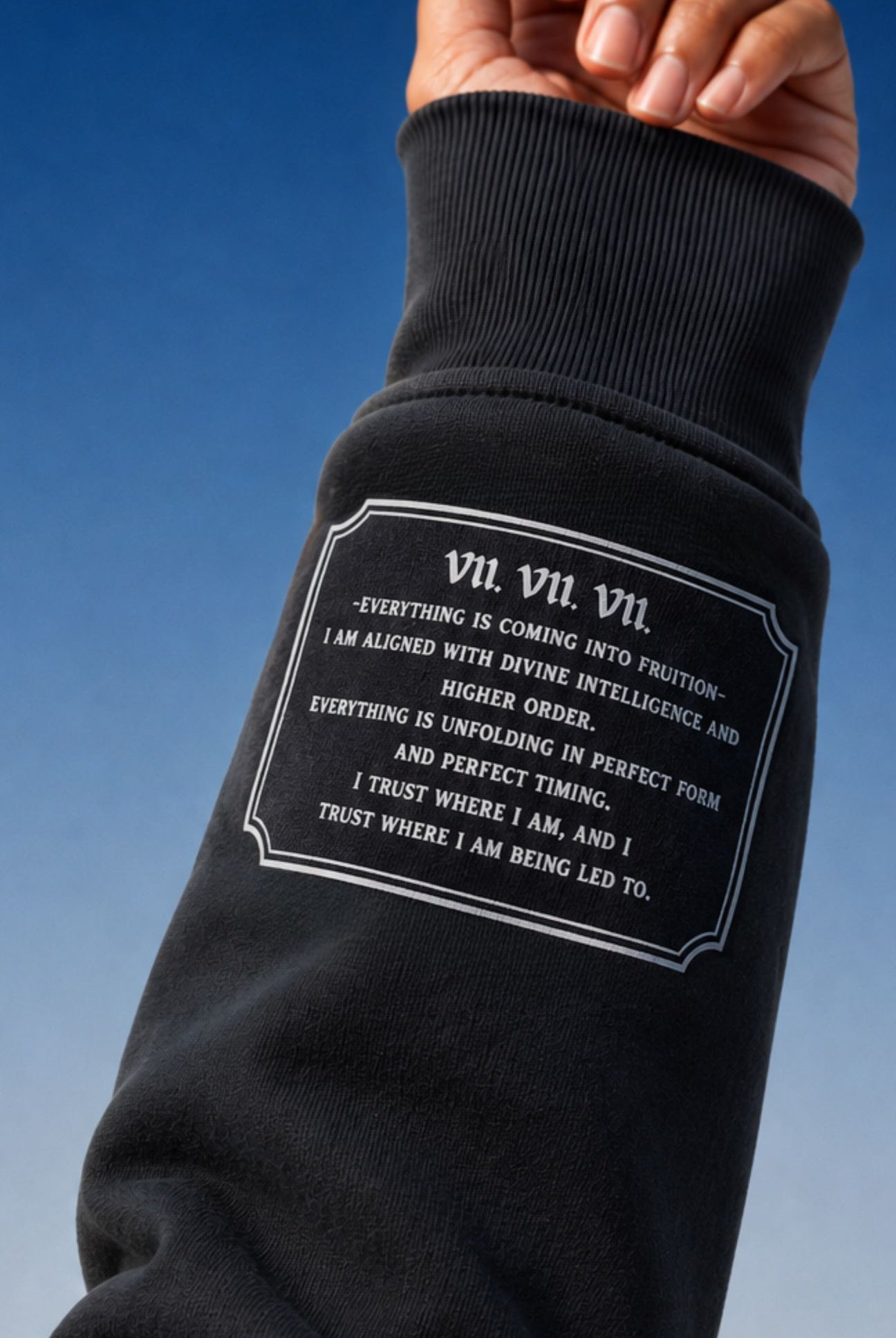 A close-up of a person's forearm wearing a black ribbed sweatshirt sleeve with a white printed quote on the front, set against a clear blue sky.