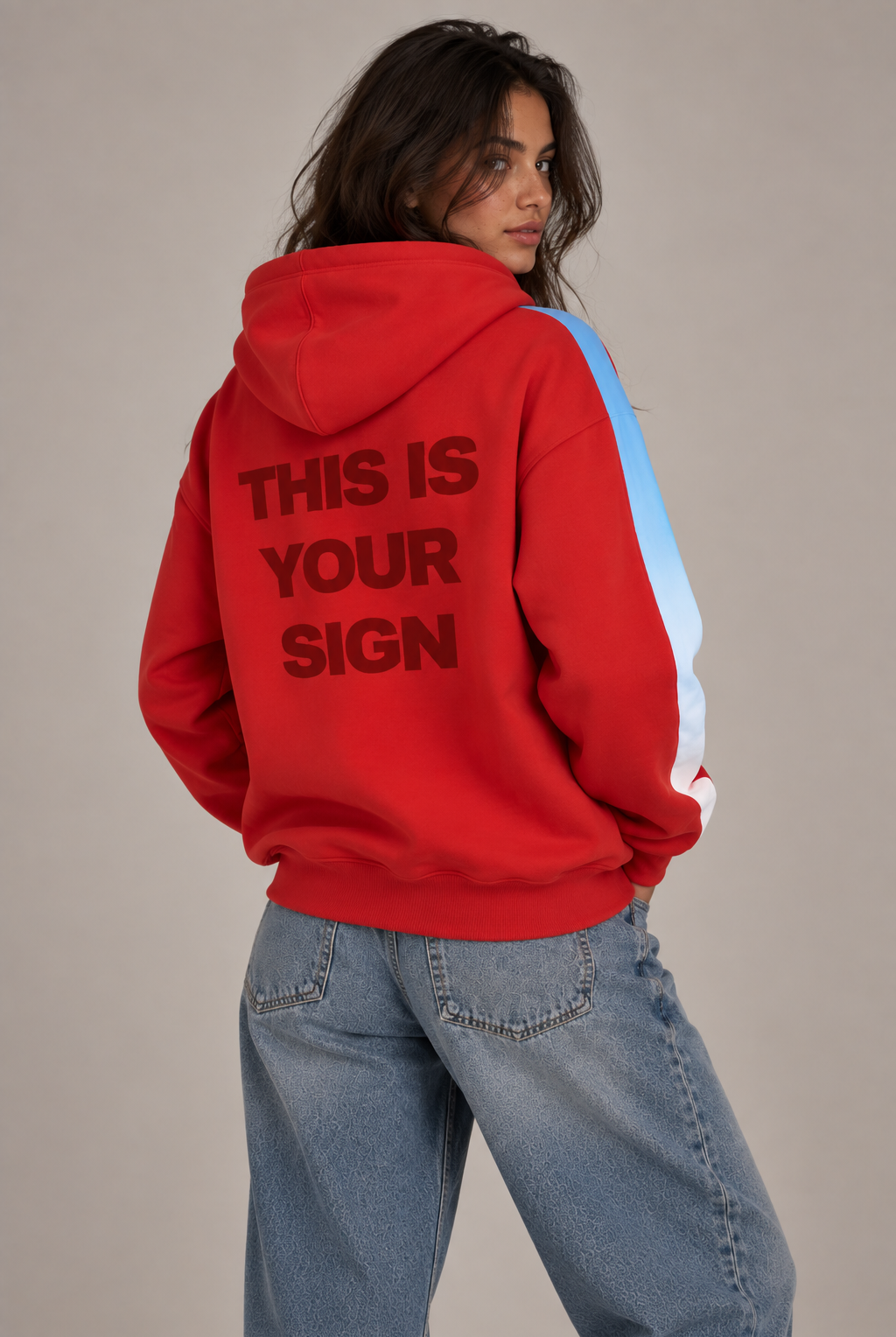 A model wearing a bright red hoodie with 'THIS IS YOUR SIGN' printed in dark redon the back, featuring light blue to white ombre detail on the sleeves, paired with light-wash blue jeans against a neutral beige background.