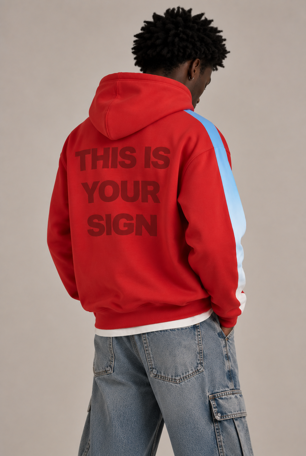 A model from behind wearing a red hoodie with 'THIS IS YOUR SIGN' printed in dark red on the back, featuring light blue and white side stripe, paired with light-wash blue jeans against a solid beige background.