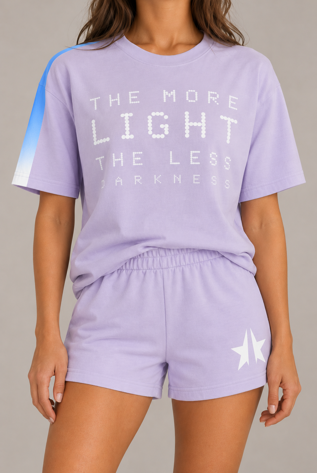 A model wearing a lavender purple matching lounge set consisting of a short-sleeve graphic t-shirt and matching shorts. The t-shirt features white text reading 'THE MORE LIGHT THE LESS DARKNESS' and a blue-to-white gradient stripe on the right sleeve.