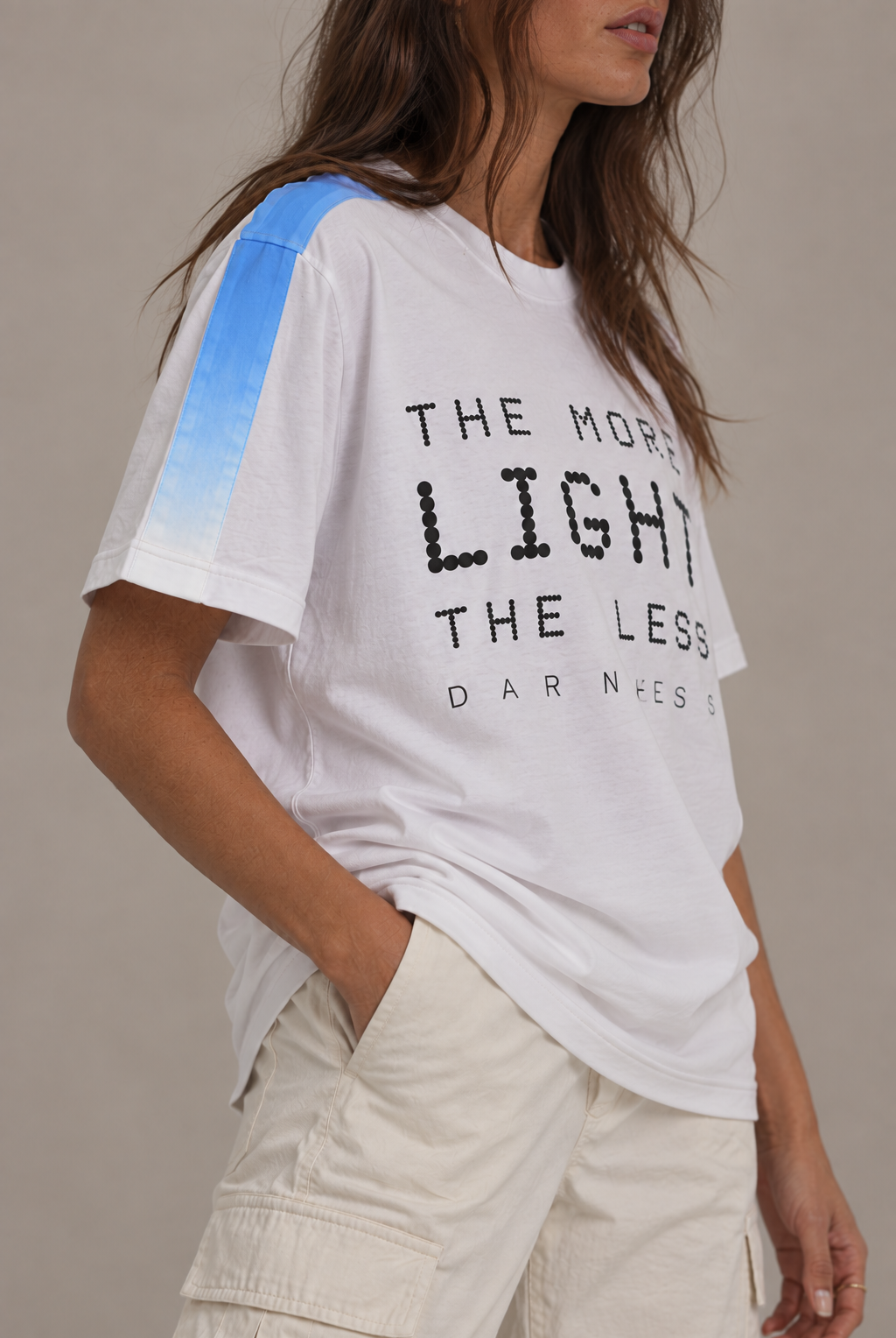 A woman wearing a white graphic t-shirt with a black text print reading 'THE MORE LIGHT THE LESS DARNNESS' and a blue gradient stripe on the shoulder, paired with beige cargo pants against a neutral background.