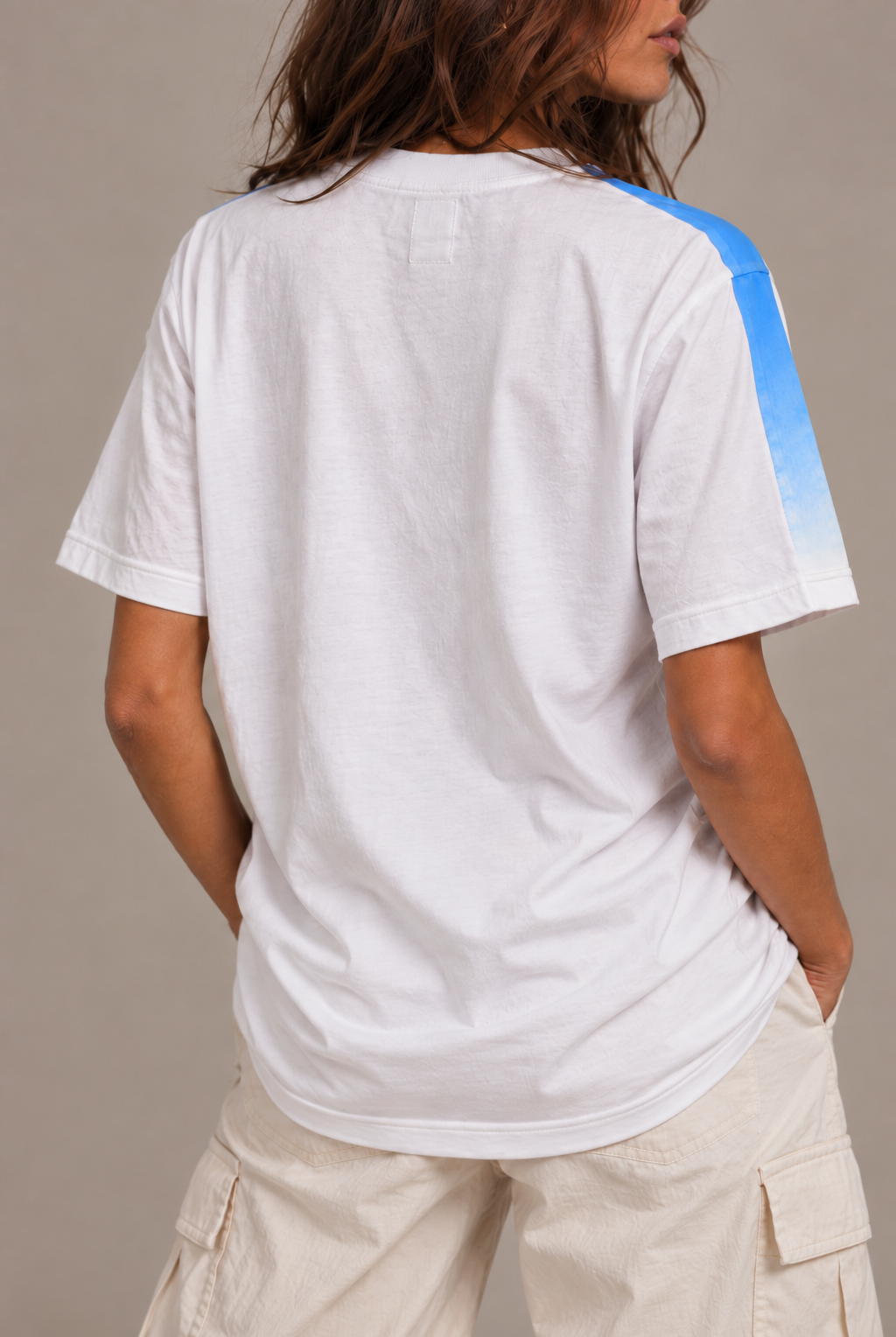 A woman wearing a white short-sleeve t-shirt with a blue gradient stripe down the right sleeve, shown from the back against a neutral studio background.