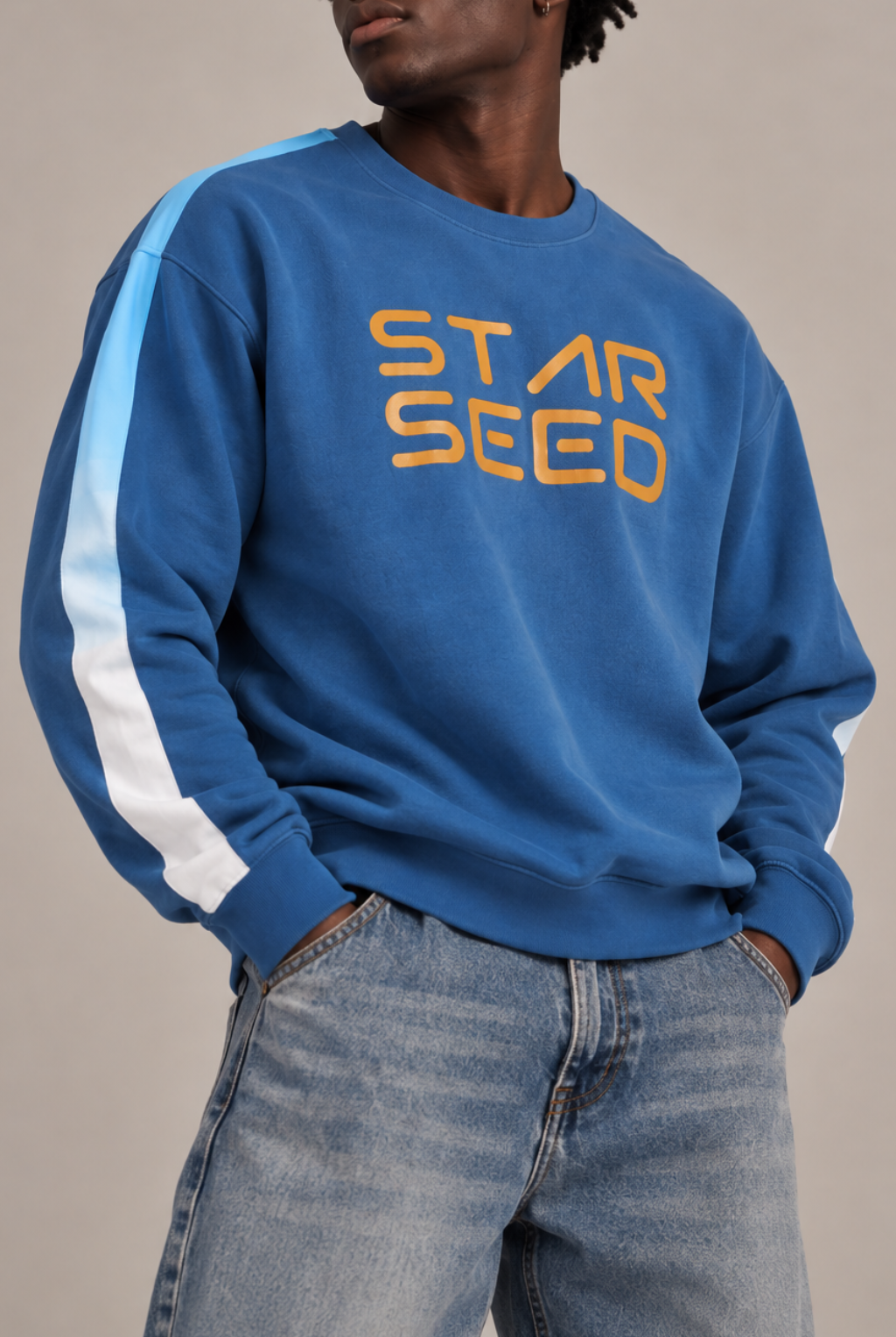 A man wearing a blue crewneck sweatshirt with 'STAR SEED' printed in orange across the chest and light blue and white stripes on the sleeve, paired with blue jeans against a neutral beige background.