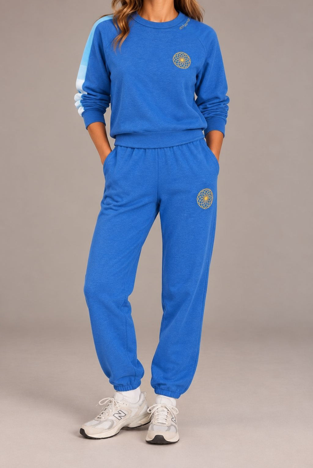 A woman modeling a matching royal blue sweatshirt and jogger sweatpants set with white sneakers, photographed against a neutral gray studio background.