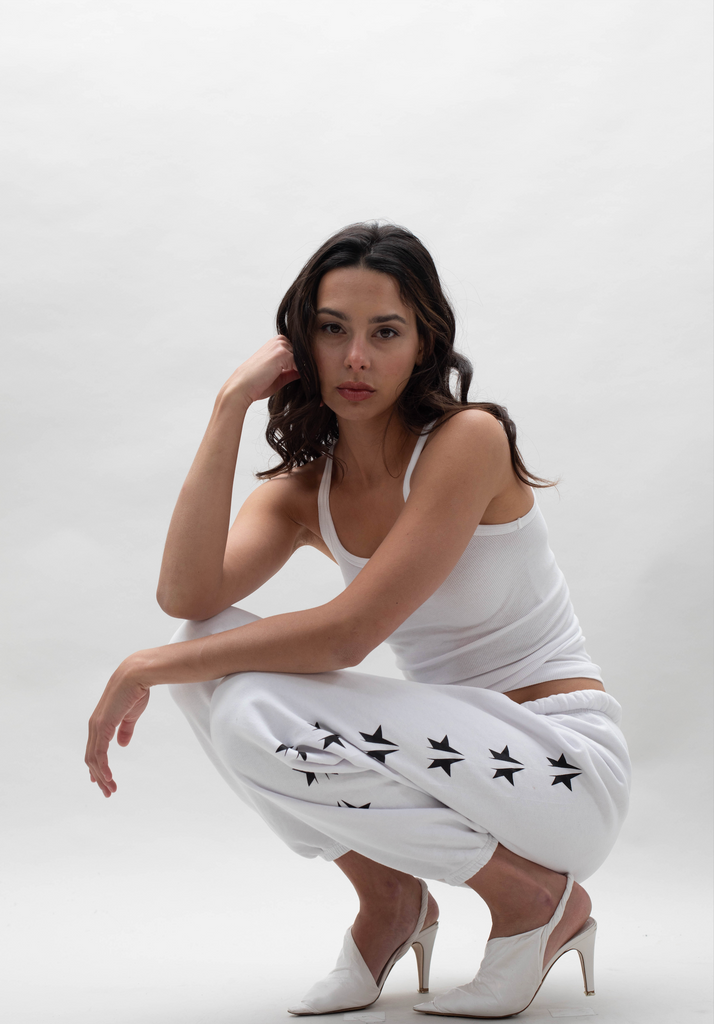 A woman squats against a plain white background. She is wearing a white tank top, white sweatpants with black stars, and white high-heeled shoes. She rests one hand on her head and looks at the camera.
