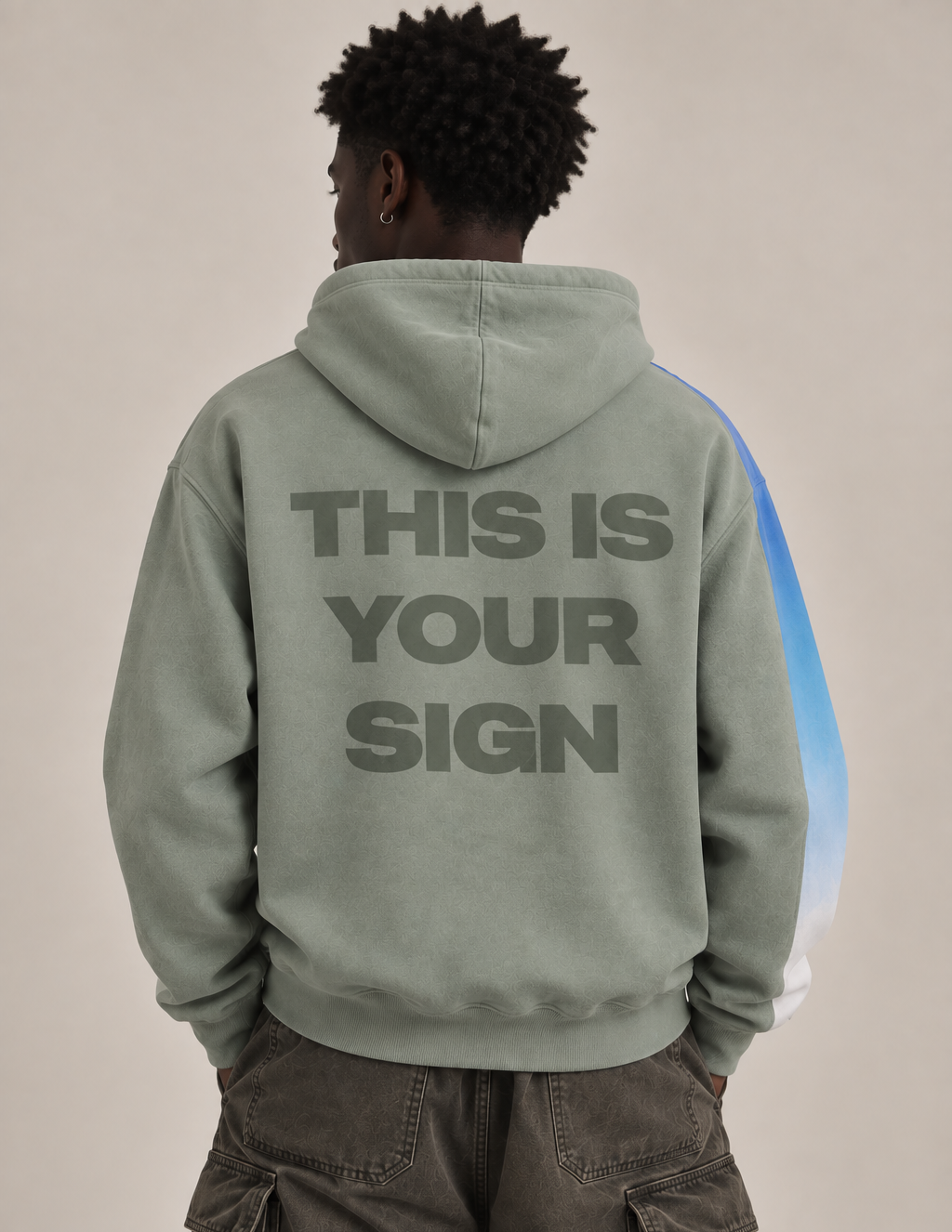 A person shown from the back wearing a sage green hoodie with the text 'THIS IS YOUR SIGN' printed in dark gray across the back, paired with dark cargo pants against a plain beige studio background.