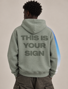 A person shown from the back wearing a sage green hoodie with the text 'THIS IS YOUR SIGN' printed in dark gray across the back, paired with dark cargo pants against a plain beige studio background.