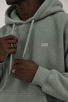 A close-up of a person wearing a sage green hooded sweatshirt with white drawstrings and a small white '1111' logo on the chest.