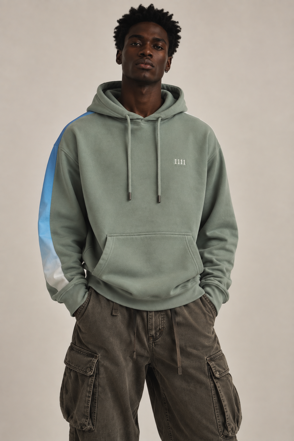 A man wearing a sage green pullover hoodie with a light blue and white gradient stripe down the sleeve, and a white embroidered '1111' on the left chest, paired with dark brown cargo pants, against a plain beige background.