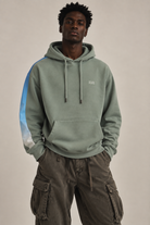 A man wearing a sage green pullover hoodie with a light blue and white gradient stripe down the sleeve, and a white embroidered '1111' on the left chest, paired with dark brown cargo pants, against a plain beige background.