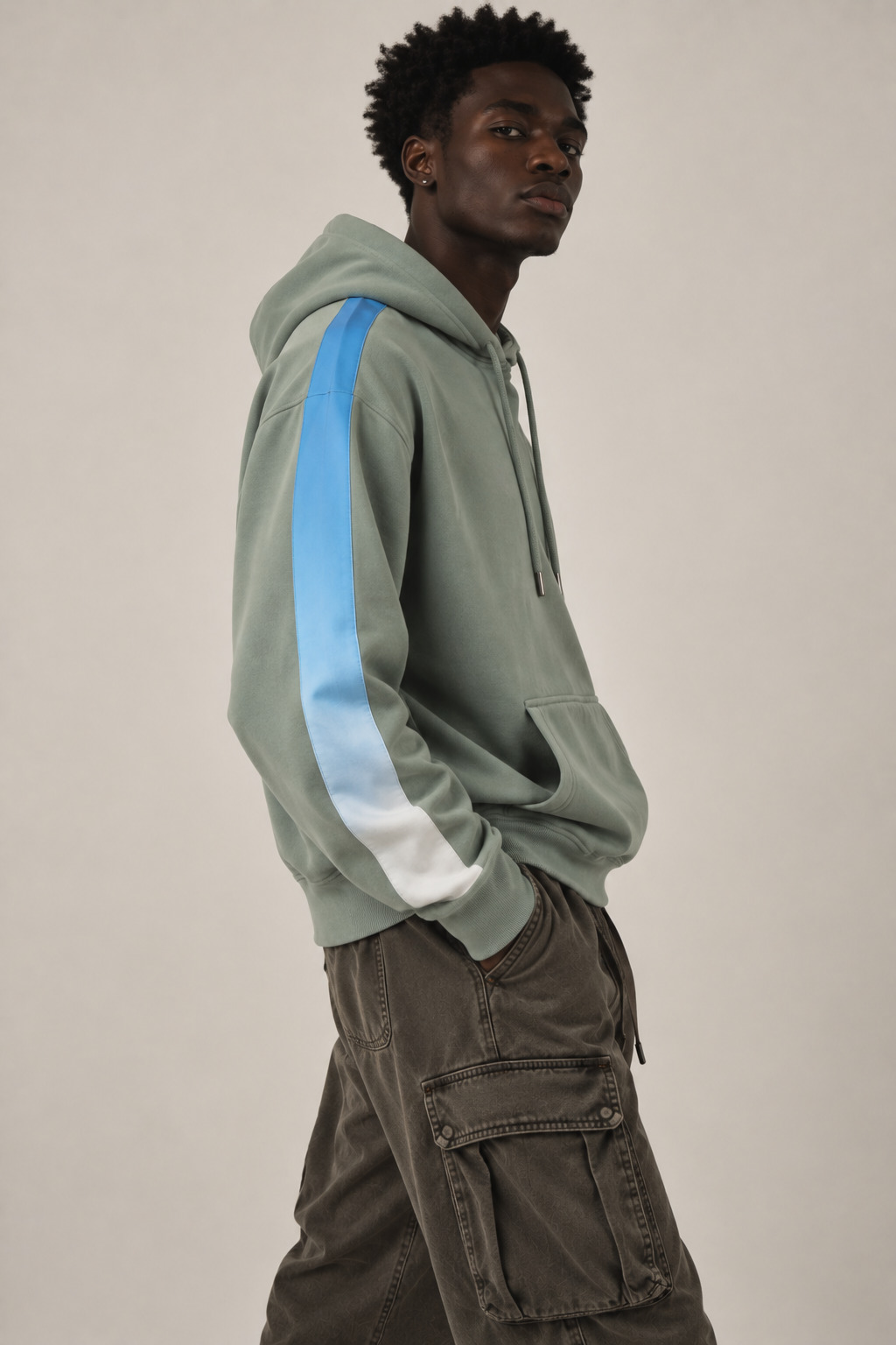 A man wearing a sage green hoodie with a light blue and white gradient stripe down the sleeve, paired with dark brown cargo pants, photographed against a neutral light gray background.