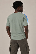 A man viewed from behind wearing a sage green t-shirt with a blue and white color-blocked stripe on the right shoulder, paired with brown cargo pants, against a plain beige background.