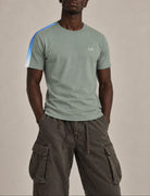 A man wearing a sage green crewneck t-shirt with a blue and white diagonal stripe on the right sleeve and a small white embroidered 1111 on the left chest, paired with dark brown cargo pants, against a solid beige studio background.