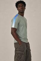 Side view of male model wearing a sage green crewneck t-shirt with white embroidered 1111 angel number detail and a blue-to-white gradient stripe along the right shoulder and sleeve, paired with dark gray cargo pants, photographed against a neutral beige studio background.