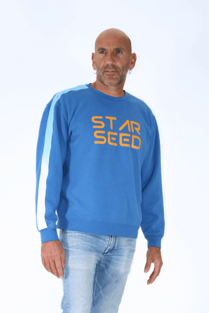 Bald man in a blue GFLApparel Starseed Crewneck sweatshirt and jeans standing against a white background, embodying his celestial heritage.