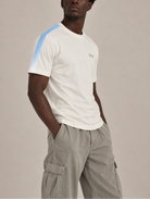 A man wearing a white short-sleeve t-shirt with a light blue stripe detail on the right shoulder and a small green '222' embroidery on the chest, paired with light gray cargo pants, against a solid light gray background.