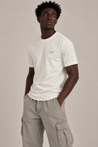 A man wearing a white crew-neck t-shirt with a small green '222' graphic on the left chest, paired with light gray cargo pants, against a solid light gray background.