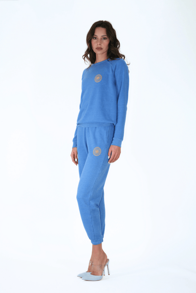 Woman posing in a blue tracksuit and high heels on a white background, holding a representation of the Lotus of Life Women's Crewneck by GFLApparel, symbolizing spiritual significance.