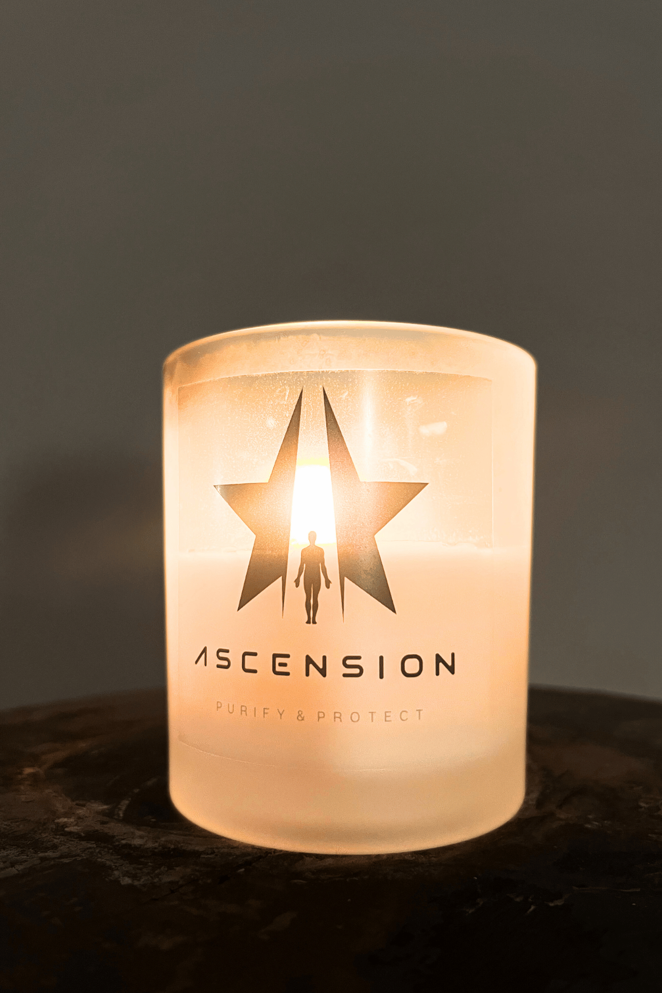 A lit Ascension Candle in a frosted glass holder features a logo with a star and figure, with "Ascension" and "Purify & Protect" printed below. Made from non-carcinogenic soy wax infused with essential oils, it promises a pure and protective ambiance. The Ascension Candle by Galactic Federation of Light offers this experience.