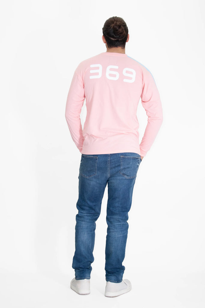 A person stands facing away, wearing a pink 369 L/S in Sunfade from GFLApparel with the number "369" on the back, blue jeans, and white shoes.