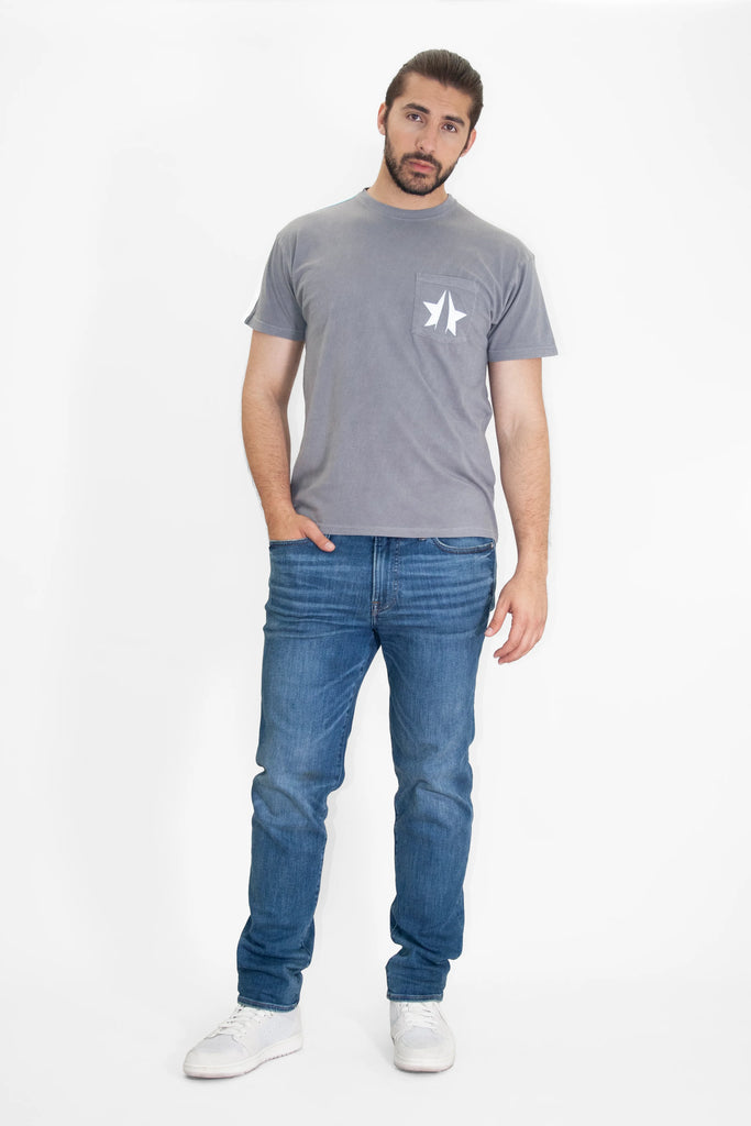 A man stands against a white background, wearing a gray cotton jersey knit 369 Tee in Asteroid by GFLApparel, blue jeans, and white sneakers. He has one hand in his pocket and looks directly at the camera.