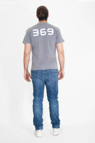 A person with long hair pulled back is standing with their back to the camera, wearing a gray 369 Tee in Asteroid by GFLApparel made from cotton jersey knit, blue jeans, and white shoes.