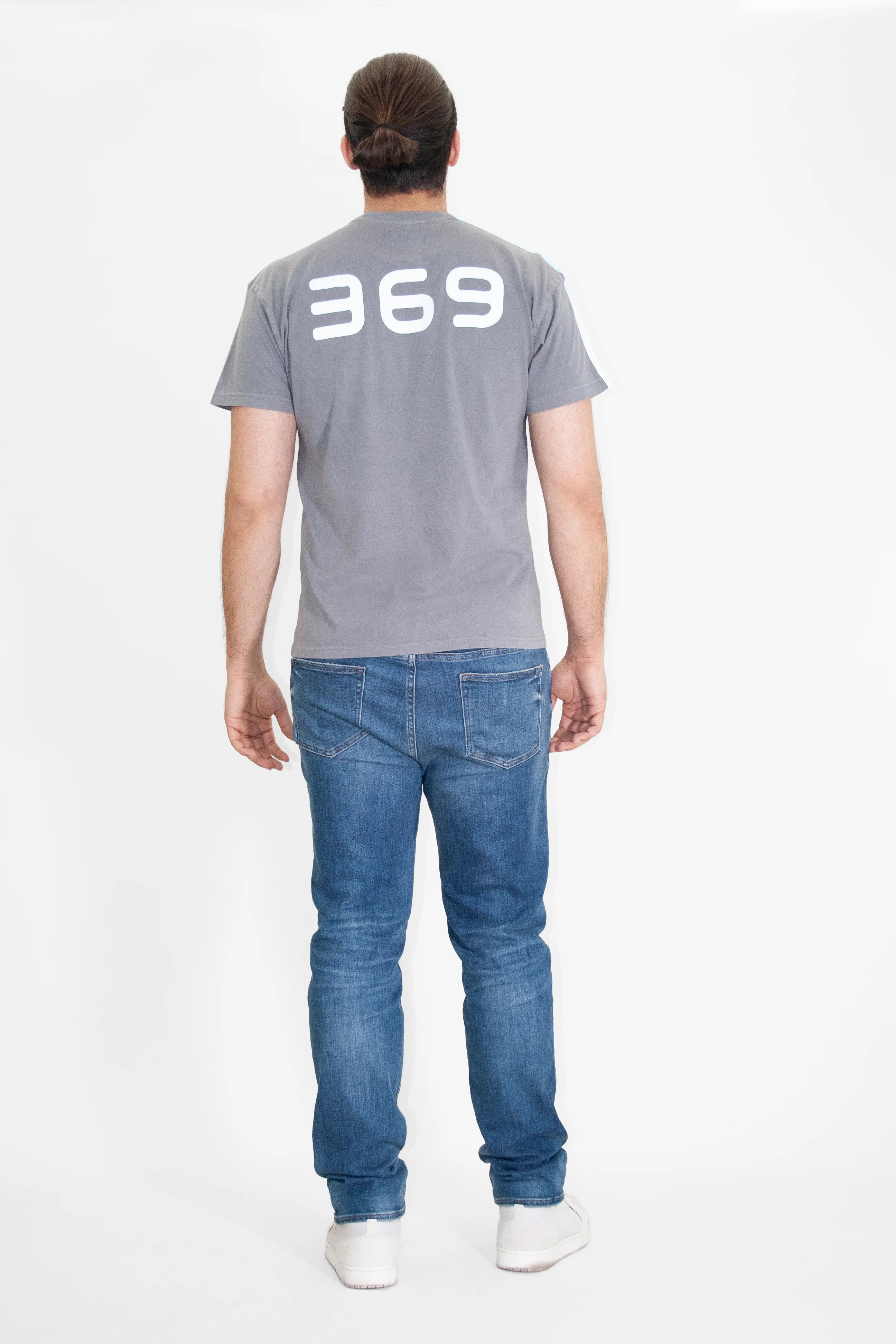 A person with long hair pulled back is standing with their back to the camera, wearing a gray 369 Tee in Asteroid by GFLApparel made from cotton jersey knit, blue jeans, and white shoes.